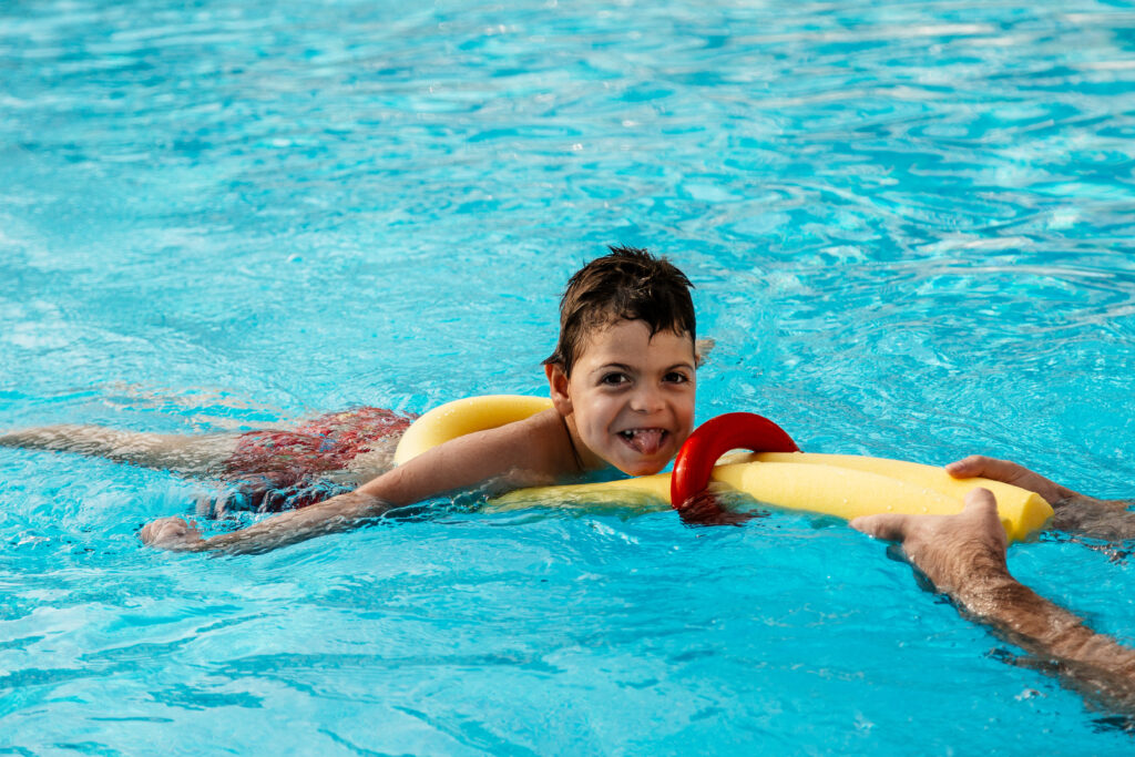 A young disabled boy is in a swimming pool. He is being supported by a yellow noodle float which has been looped under his arms and is being pulled along by an adult off the edge of the picture. The boy is sticking his tongue our at the camera.
