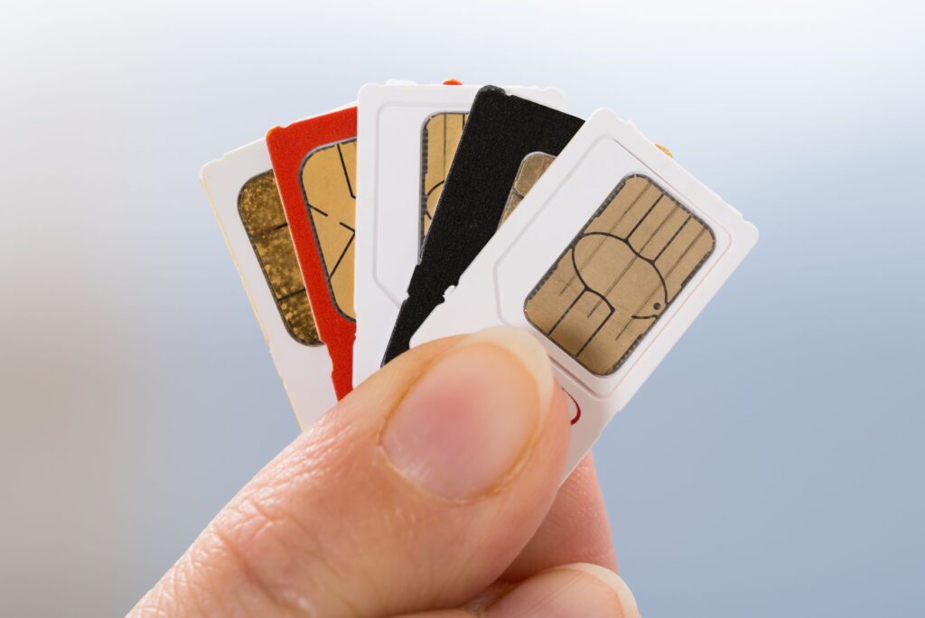 Fingers hold 5 SIM cards fanned out. The first, third and fifth SIM cards in the fan are all white. The second is red and the fourth is black.