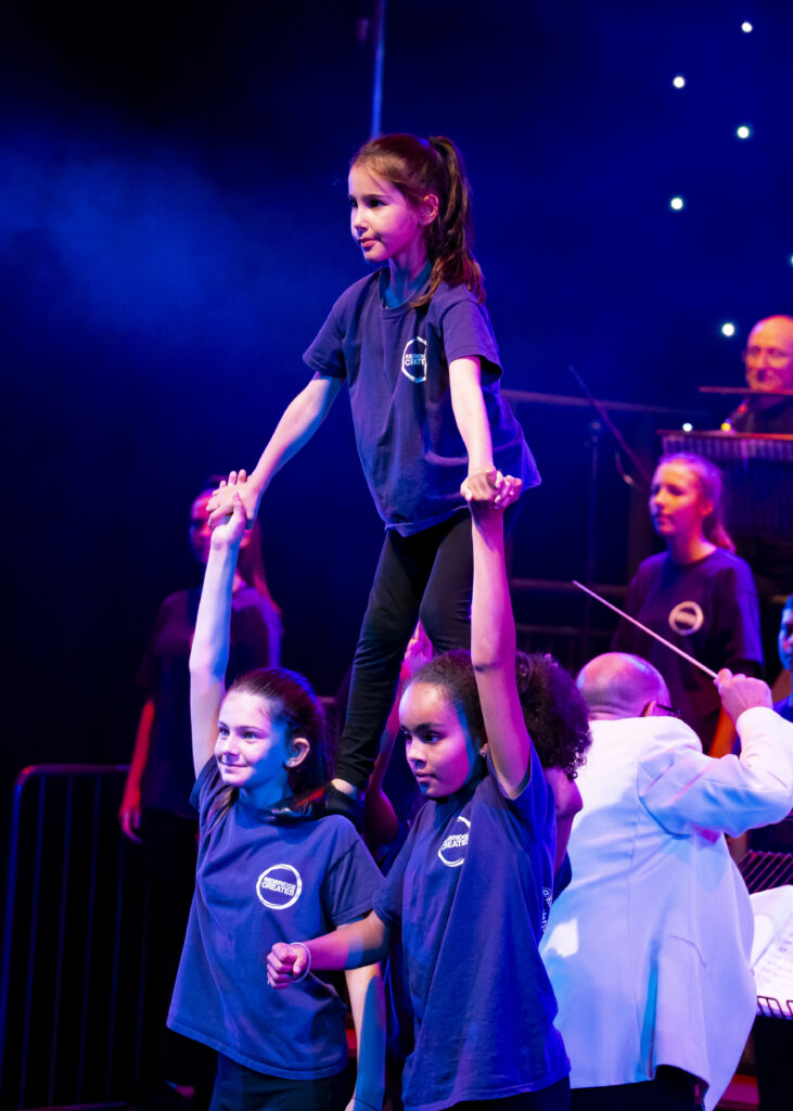 Three young girl performer standing side by side with another girl performer standing on their shoulders