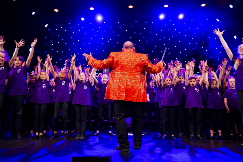 Conductor Rob Hyman on stage with a group of singers at Kenneth More Theatre. He has his back to the camera and is wearing black trousers and an orange suit jacket. Students are facing the camera and wearing purple tshirts and have their hands in the air.