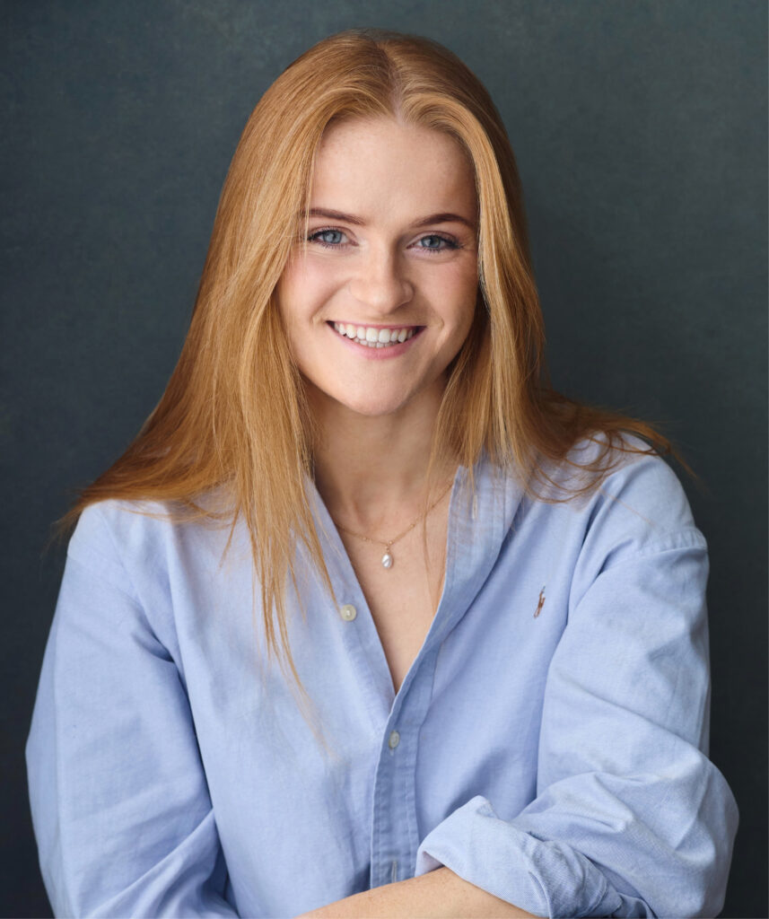 A headshot of Lydia Hunt, a white female with medium length strawberry blonde hair. She is smiling at the camera and is wearing a blue button down shit